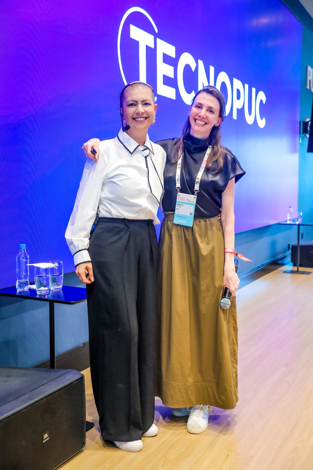 Duas idealizadoras do movimento em foto conjunta ao lado do painel Tecnopuc.