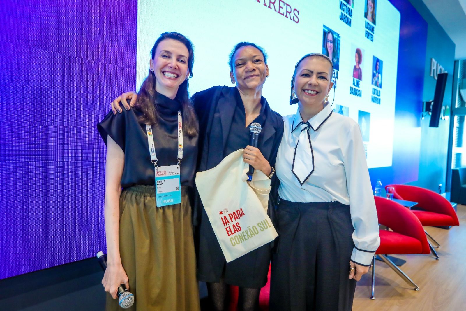 Três participantes sorriem juntas no palco segurando a ecobag do IA para Elas: Conexão Sul.
