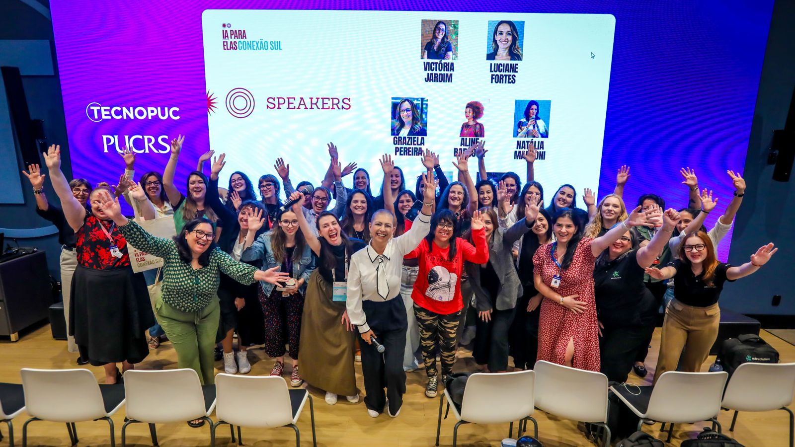 Participantes do primeiro encontro reunidas no palco com os braços erguidos, ao lado do painel de speakers.