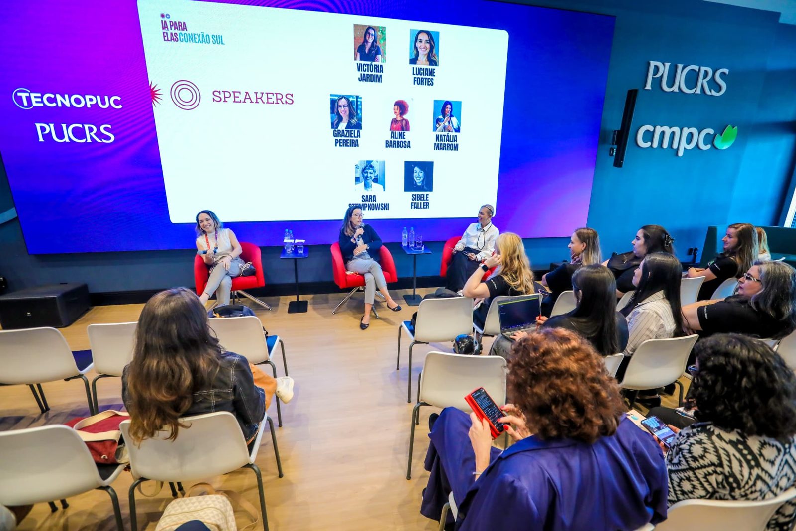 Painel de conversa no palco com o slide de speakers do IA para Elas: Conexão Sul ao fundo.
