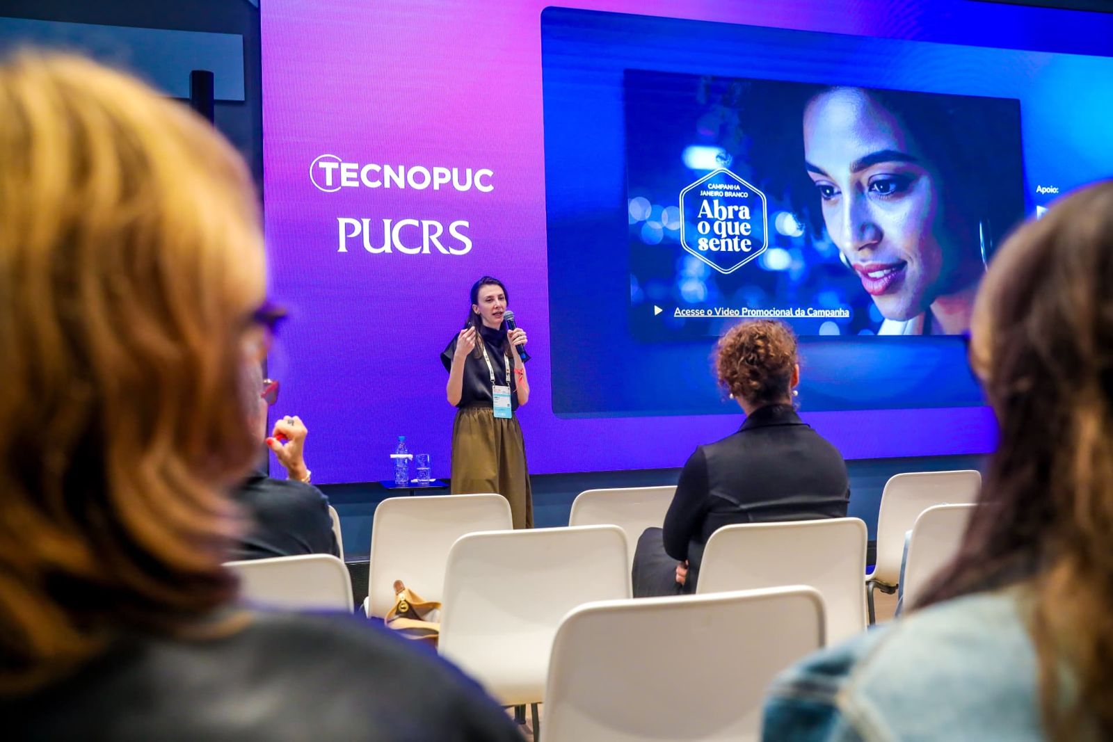 Palestrante no palco durante apresentação, com os painéis do Tecnopuc e da PUCRS ao fundo.